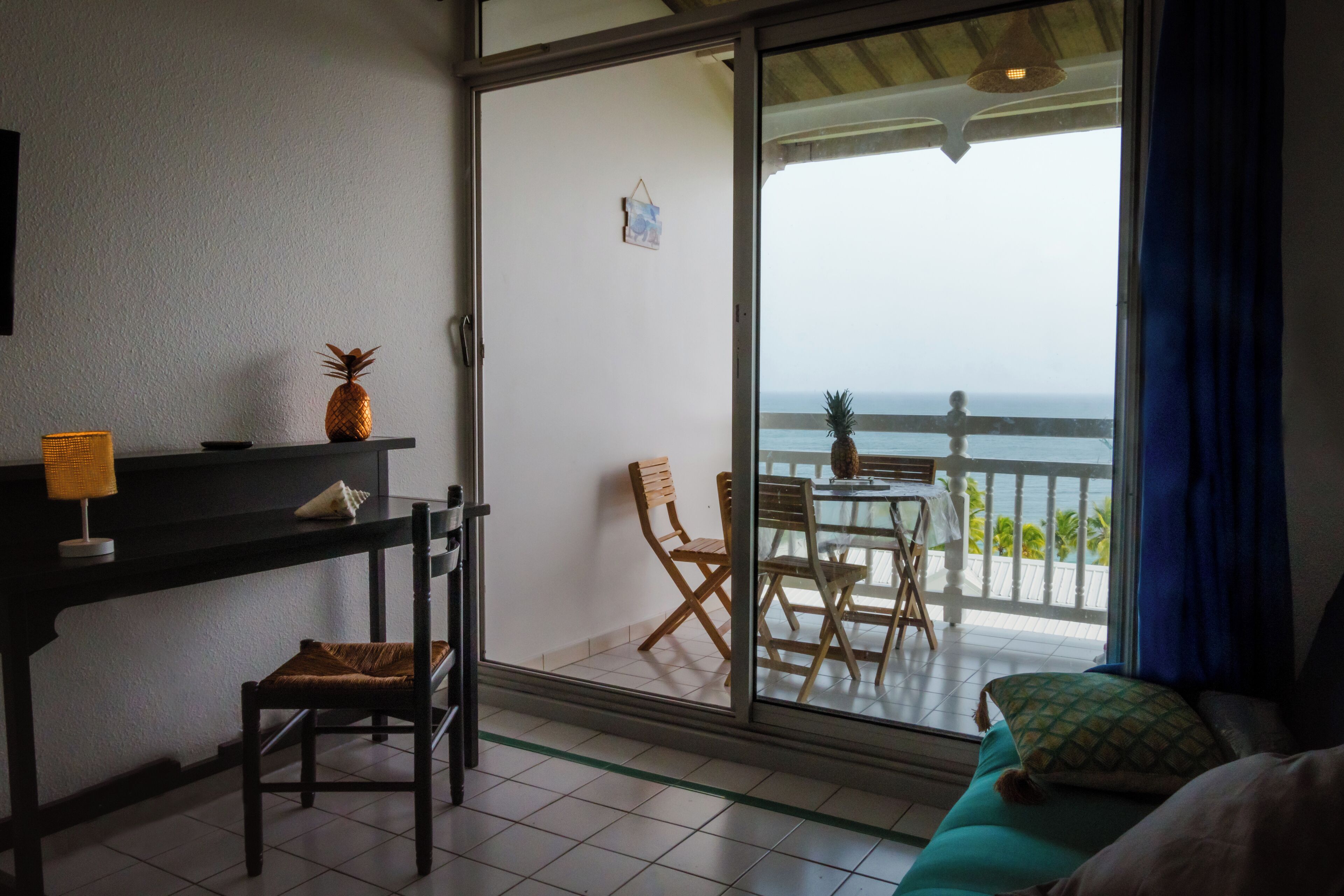 A room with a sliding glass door leading to a balcony with a table and chairs, a bed, a desk with a lamp, and a pineapple on the desk.