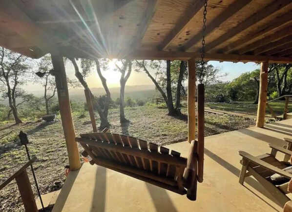 Une balançoire en bois, placée sous une zone abritée, d’où l’on peut admirer les arbres et un paysage lointain.