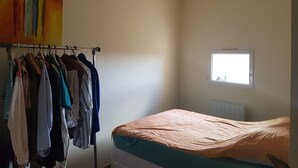 A bedroom with a bed, a window, a radiator, and a clothing rack with various garments. - Location Maison de Village, vue Exceptionnelle (Grambois)