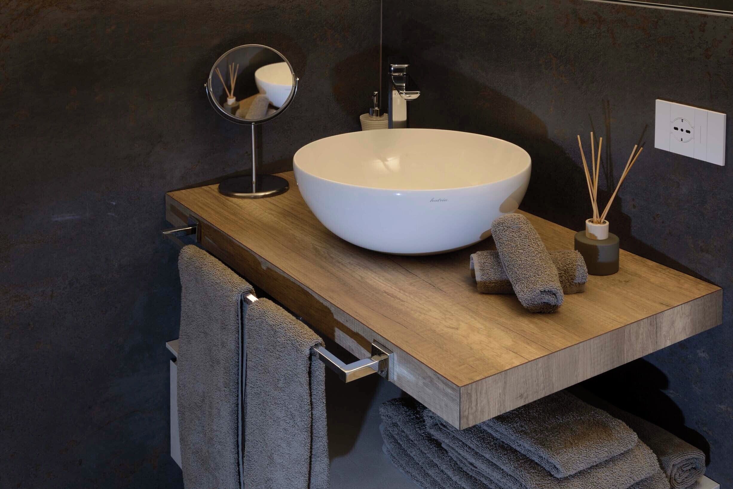 A modern bathroom with a large white vessel sink, a wooden vanity, a towel rack, a mirror, and a small container with reed sticks.