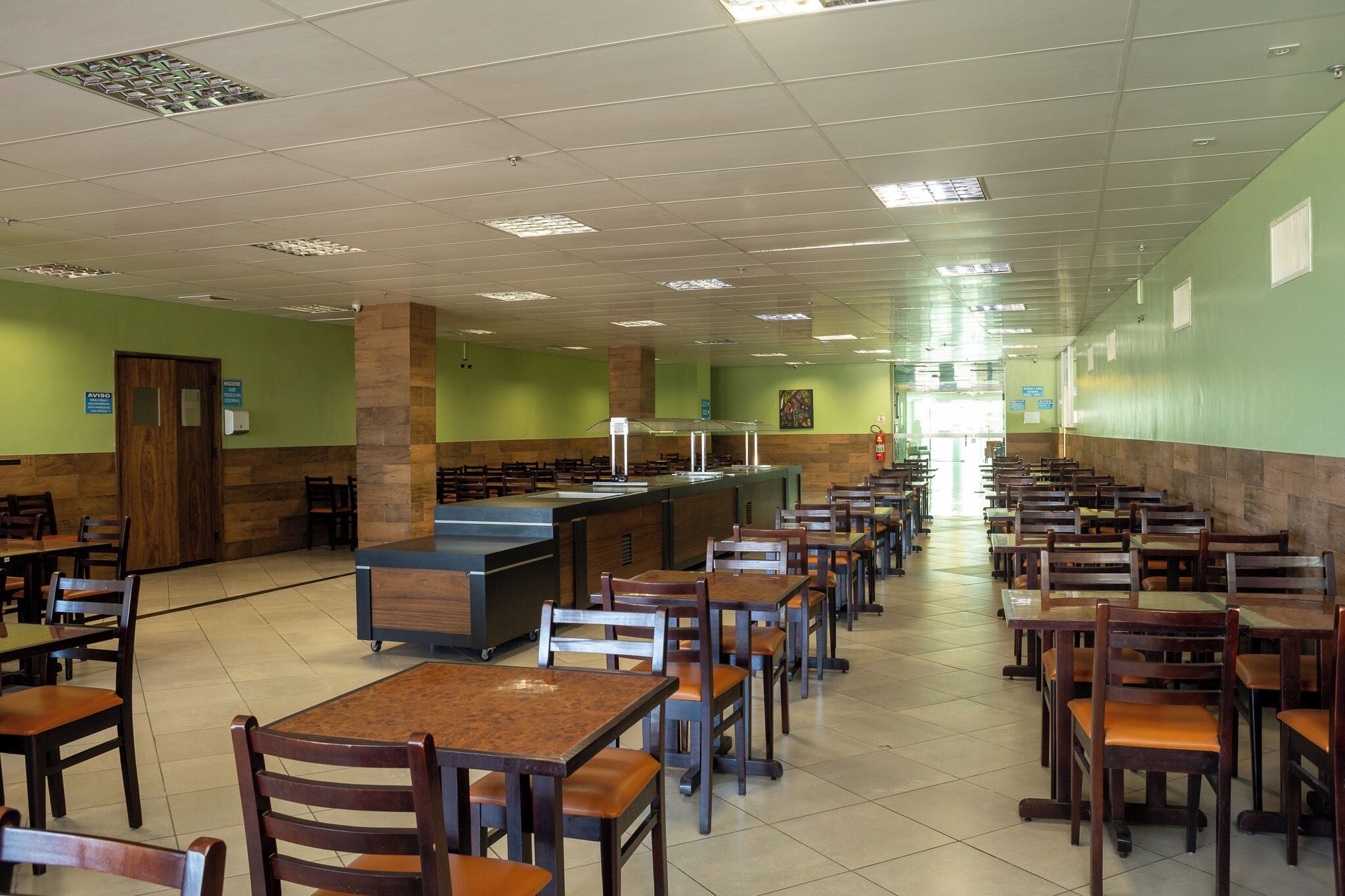 A cafeteria with long tables and chairs, a central serving counter, and green walls.