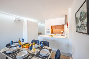 A modern dining area with a kitchen in the background, a dining table set with plates, glasses, and a fruit basket.