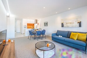A modern living room with a blue sofa, a glass coffee table, and a dining area with blue chairs.