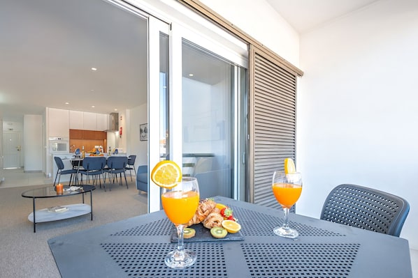 A dining area with a table set for two, featuring a glass of orange juice and croissants, and a view of a modern kitchen in the background.