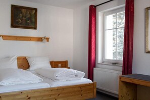 Ein Schlafzimmer mit einem hölzernen Bett, weißer Bettwäsche, einem roten Vorhang, einem Fenster mit Blick auf Bäume und einem Gemälde an der Wand.