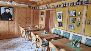 Ein Esszimmer mit Holzwandverkleidung, einem langen Tisch mit Stühlen, einer Sitzbank an der Wand und verschiedenen Küchenutensilien auf Regalen.