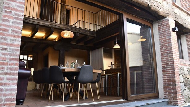 An outdoor dining area with a brick wall, wooden beams, and a glass door leading to a balcony. - La Casa del Guarda, an exclusive rural house 4* (Sotosalbos)