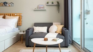 Une chambre moderne avec un lit, un canapé gris, une petite table blanche et une fenêtre donnant sur un beau paysage.