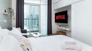 Une chambre d’hôtel avec un grand lit, une petite table, un téléviseur affichant le logo « NETFLIX », un balcon donnant sur des immeubles et une étagère avec des objets décoratifs.