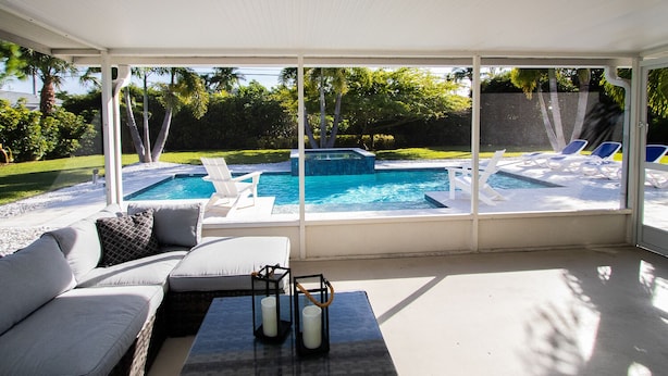 A sunroom with a pool, a sofa, and two candles on a table. - Three-bedroom home with backyard heated pool oasis. 5 minutes to the beach (Naples)