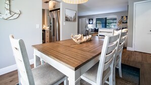 A dining area with a wooden table and white chairs, a hanging light fixture, and a kitchen in the background. - Three-bedroom home with backyard heated pool oasis. 5 minutes to the beach (Naples)