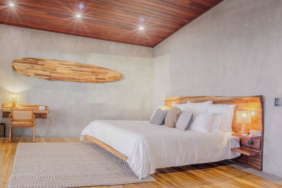 A modern bedroom with a wooden ceiling, a large bed with white bedding, a wooden headboard, a nightstand with a lamp, and a wall-mounted wooden shelf.
