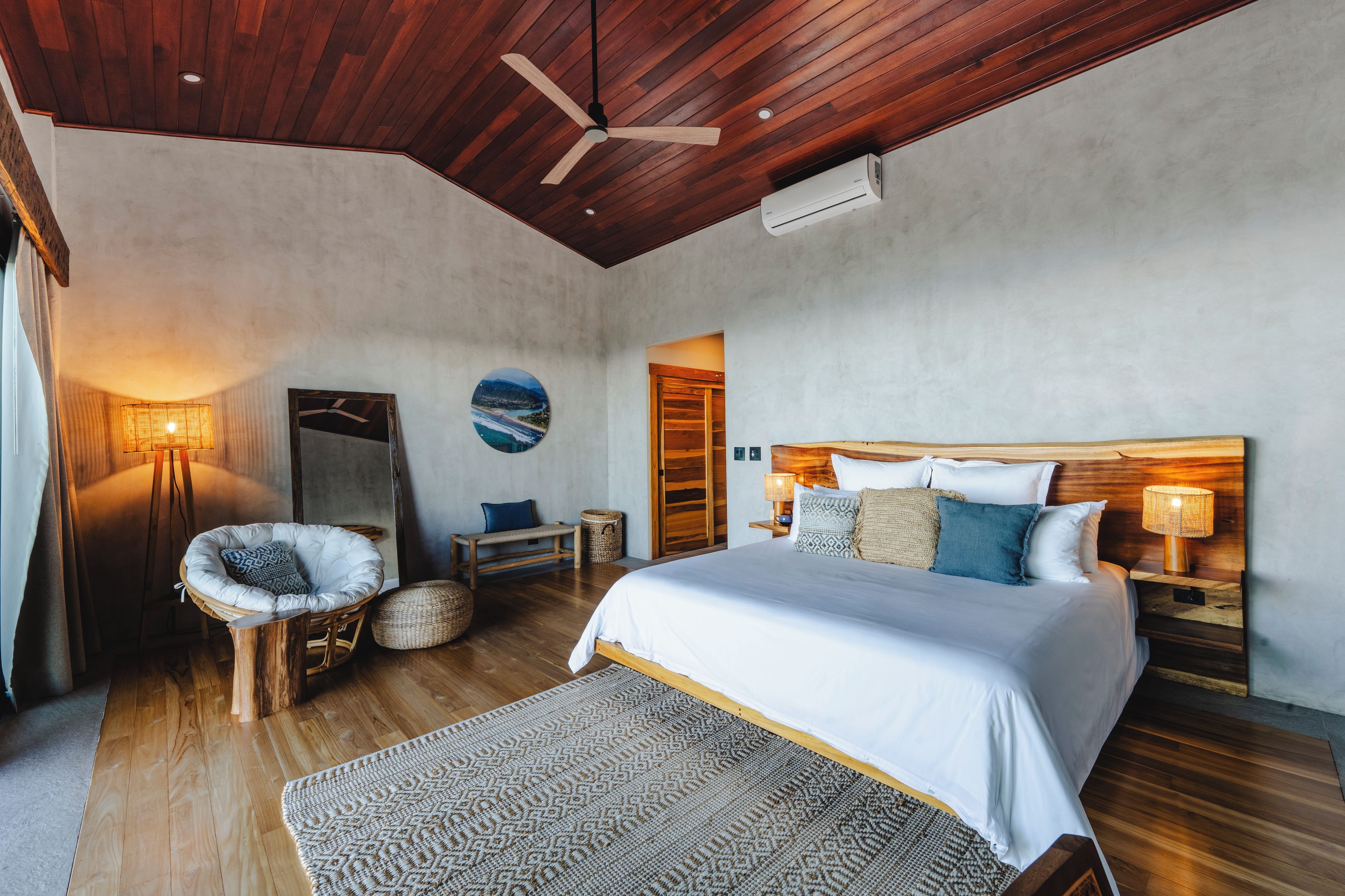 Un dormitorio con techo de madera, cama con ropa de cama blanca, espejo redondo en la pared y vista al mar.