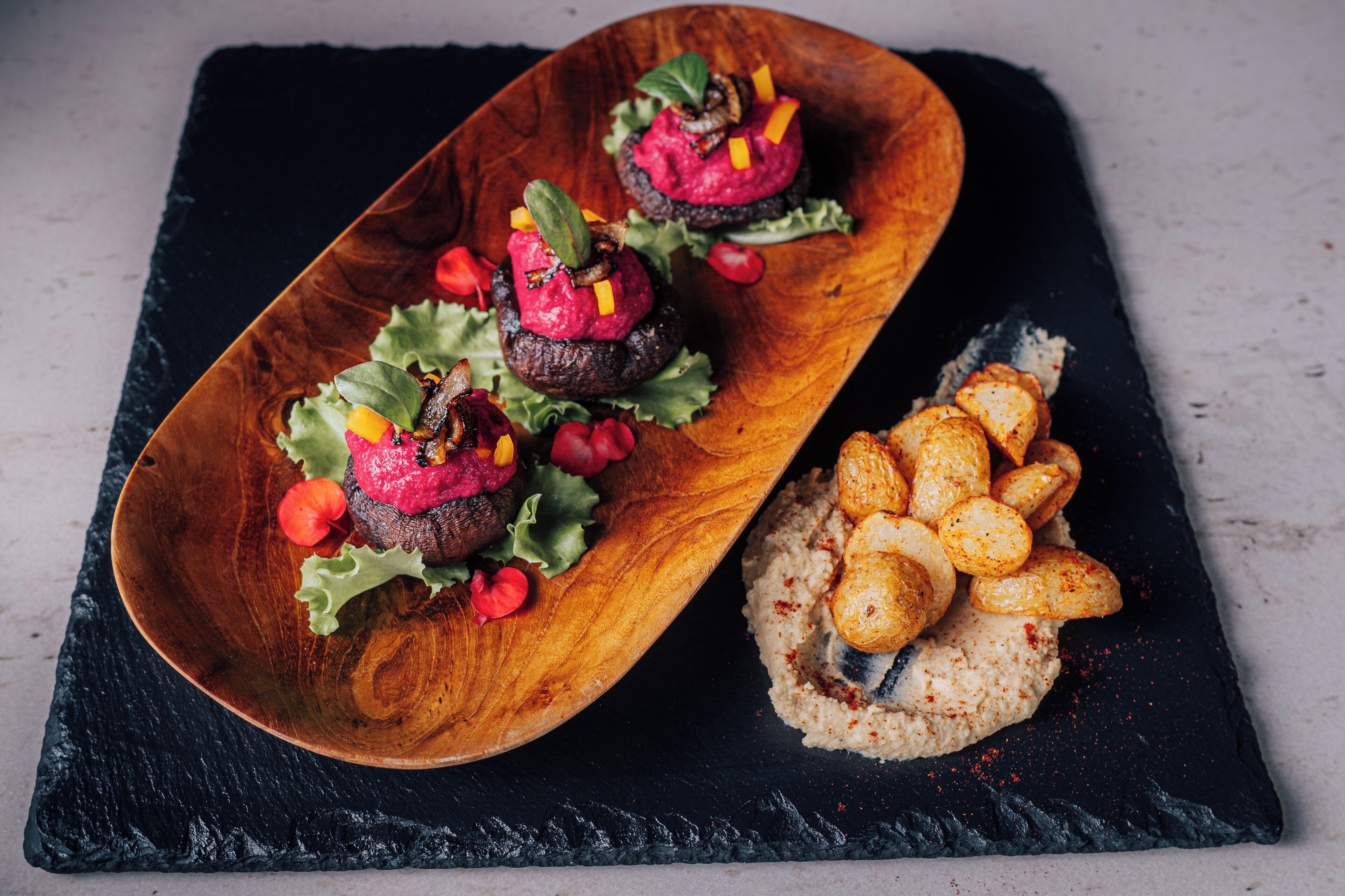 A wooden serving dish with a beetroot and mushroom dish, accompanied by roasted potatoes and a side of hummus.