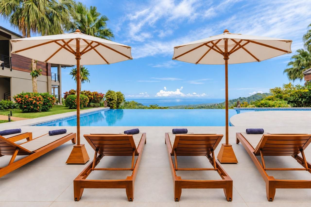 A poolside area with wooden lounge chairs and umbrellas, a clear sky, and a view of the ocean.