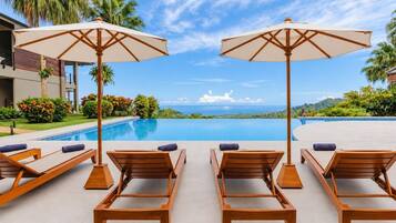 A poolside area with wooden lounge chairs and umbrellas, a clear sky, and a view of the ocean.
