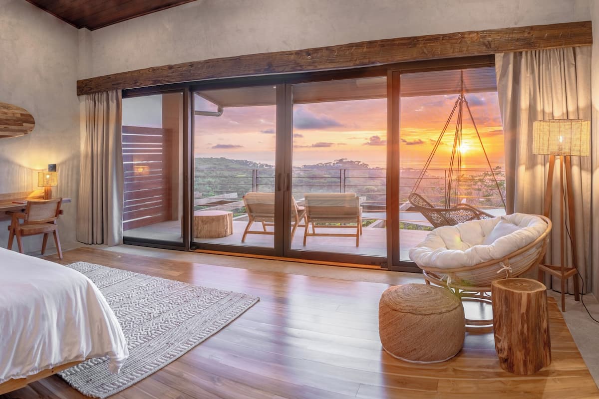 A bedroom with a large bed, a desk, and a chair, a hanging chair, and a view of the sunset through a sliding door.