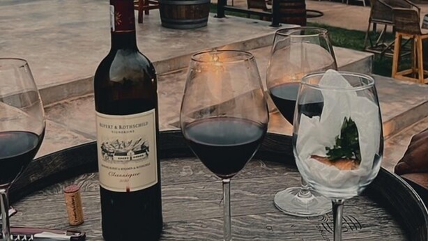 A wine tasting setup with a bottle and glasses on a wooden table, outdoor seating, and a building with a metal roof in the background. - The Shed Venue & Boutique Hotel (Chitedze)