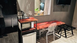 A compact hotel room with a kitchenette, a small refrigerator, a wooden desk, a chair, a flat-screen TV, and a mirror reflecting the room. - The Shed Venue & Boutique Hotel (Chitedze)