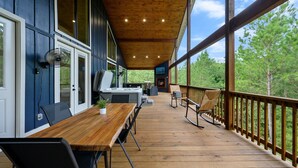Una terrazza coperta con un tavolo e sedie di legno, un barbecue e uno schermo televisivo.