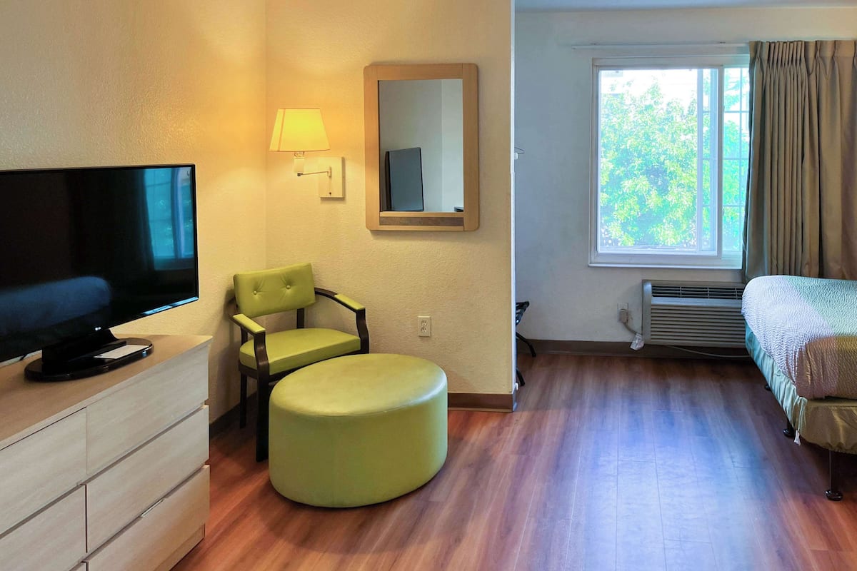A hotel room with a flat-screen TV, a green chair, a round ottoman, a bed, a window with curtains, and a mirror on the wall.