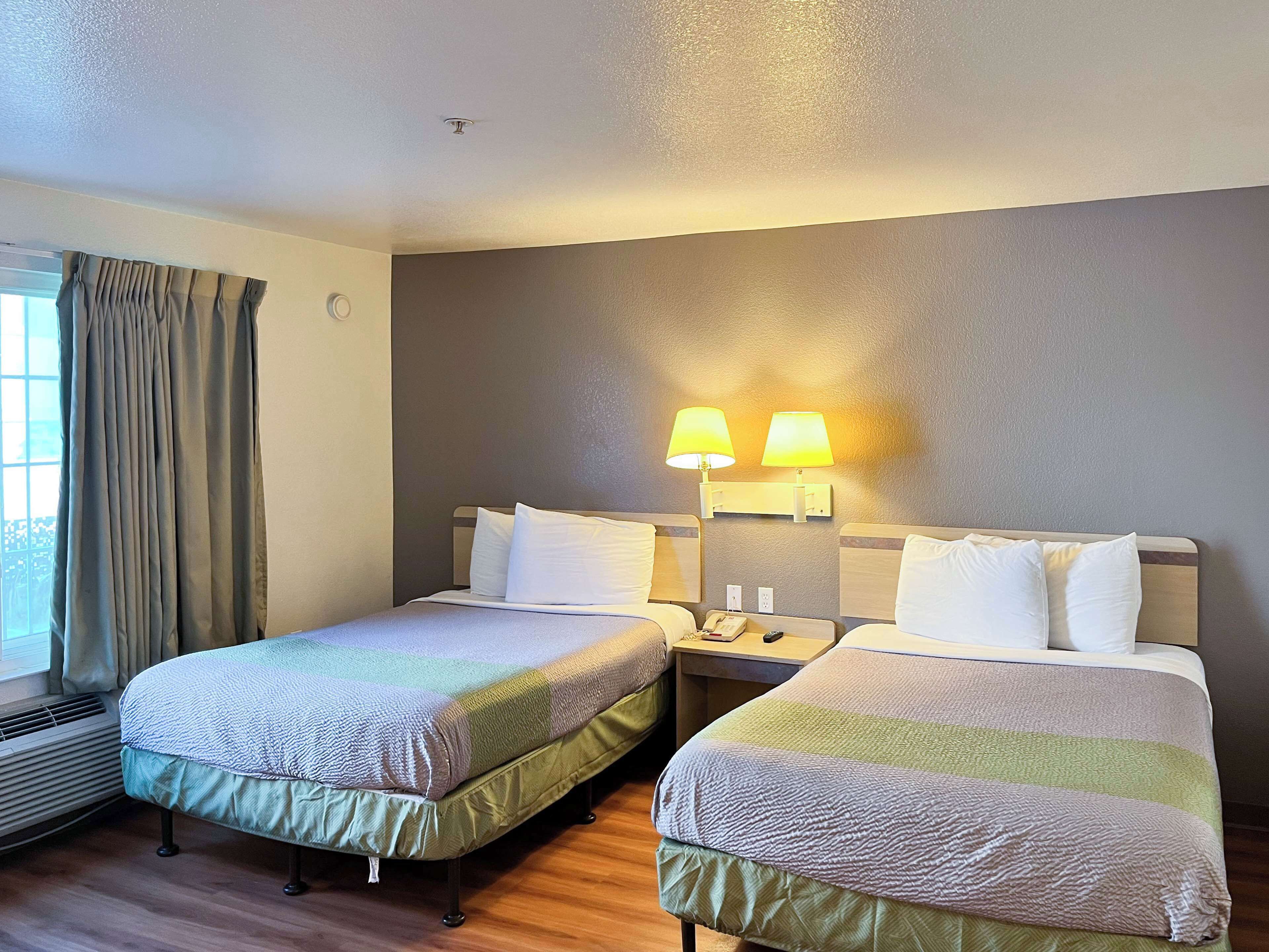 A hotel room with two beds, a window with curtains, a wall-mounted lamp, and a thermostat.