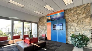 A reception area with a stone wall, a welcome sign, a digital display, and seating with potted plants. - Studio 6 Belmont, CA – San Francisco - Redwood (Belmont)