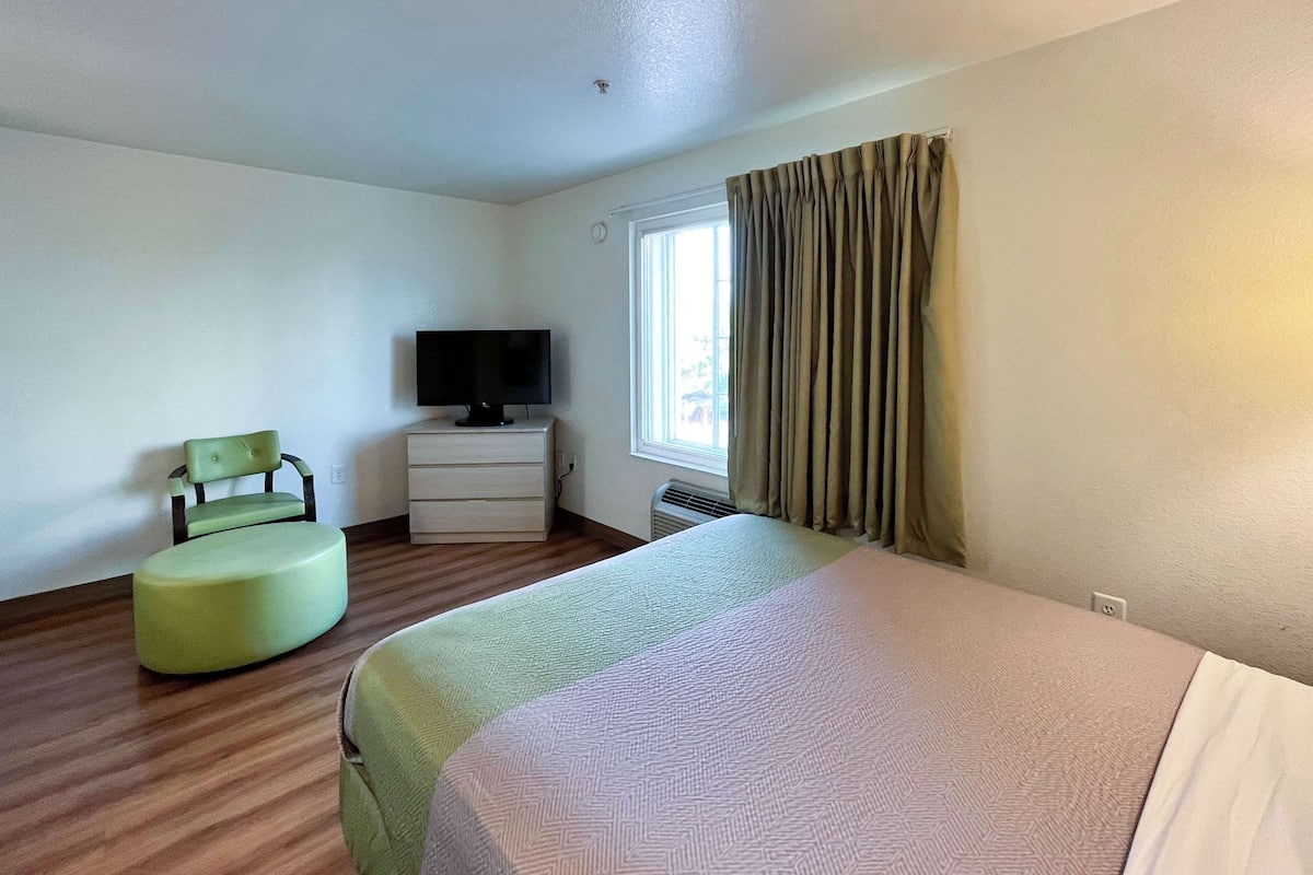 A hotel room with a bed, a green chair, a TV on a dresser, and a window with curtains.