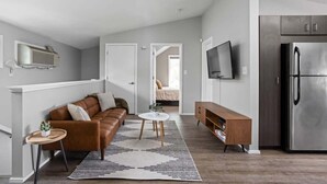 Una sala moderna con un sofá de cuero marrón, una mesa de centro redonda y un tapete a rayas. Se ve un mueble de madera y un refrigerador de acero inoxidable.