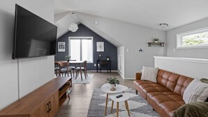 Una sala moderna con un televisor de pantalla plana grande, un sofá de cuero marrón, una zona de comedor con mesa y sillas, y una escalera que lleva a un nivel superior.