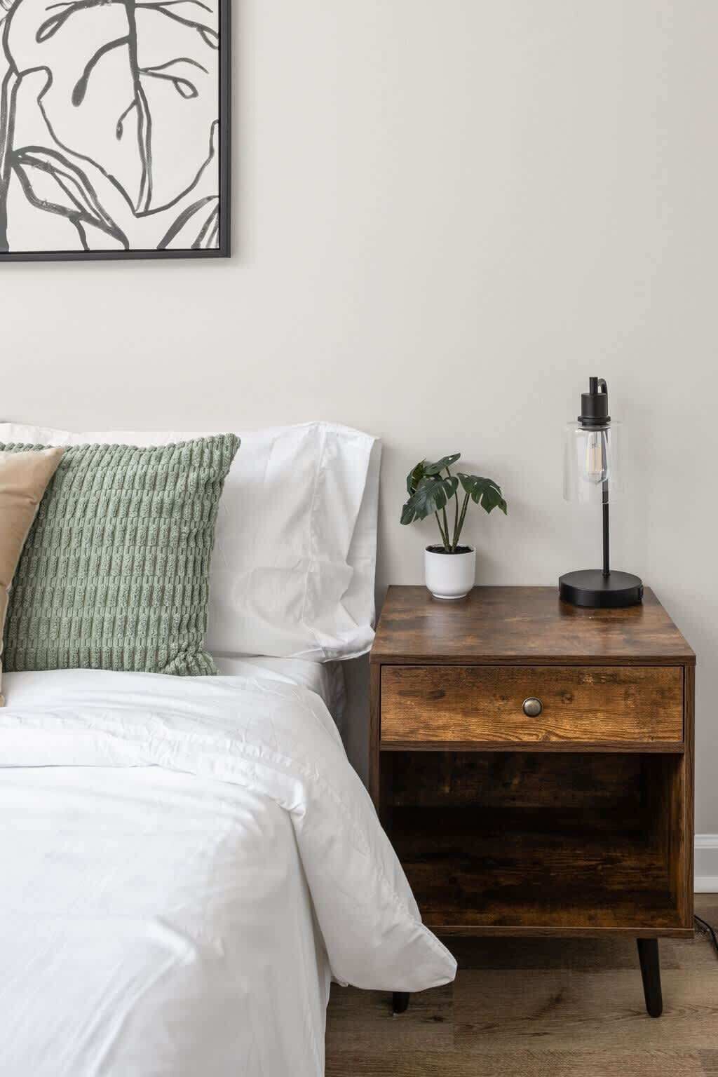 Une chambre à coucher avec un lit, une table de chevet avec une lampe, une plante en pot et une œuvre d’art encadrée accrochée au mur.