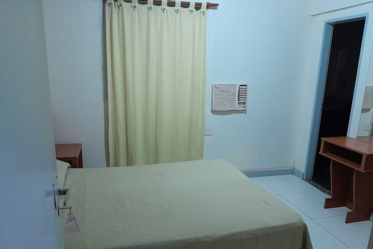 Una habitación con una cama, una cortina, aire acondicionado instalado en la pared, una puerta y una pequeña mesa.