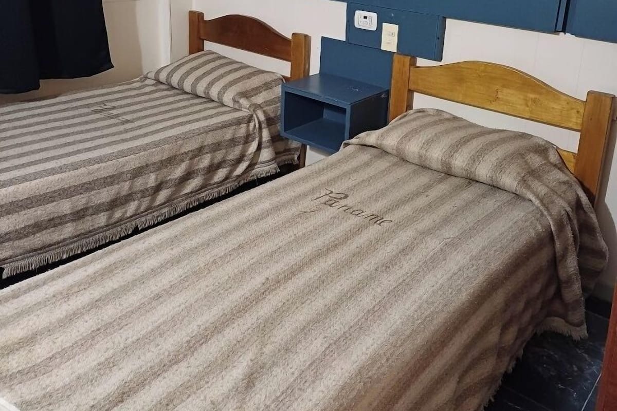 Dos camas individuales con ropa de cama a rayas, una mesita y una lámpara de pared en una habitación con paneles blancos y azules.