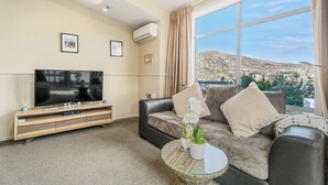 Una sala de estar con un televisor de pantalla plana grande, un sofá, una mesa de centro con una planta y vistas a un paisaje montañoso a través de un ventanal amplio.