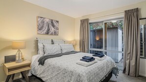 Un dormitorio con una cama grande, una mesita de noche con lámpara, una obra de arte enmarcada sobre la cama y vistas al exterior a través de una ventana.
