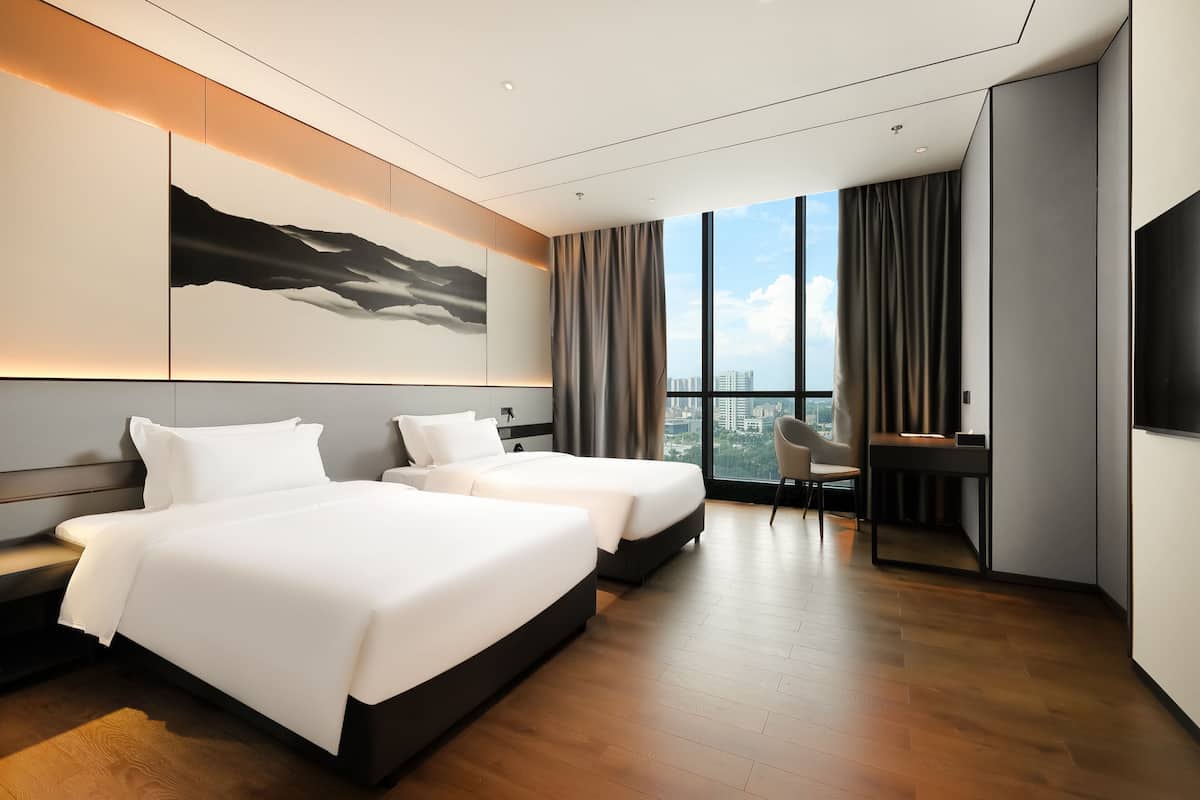 Quarto de hotel moderno com duas camas, uma escrivaninha e uma janela grande com vista para a cidade.