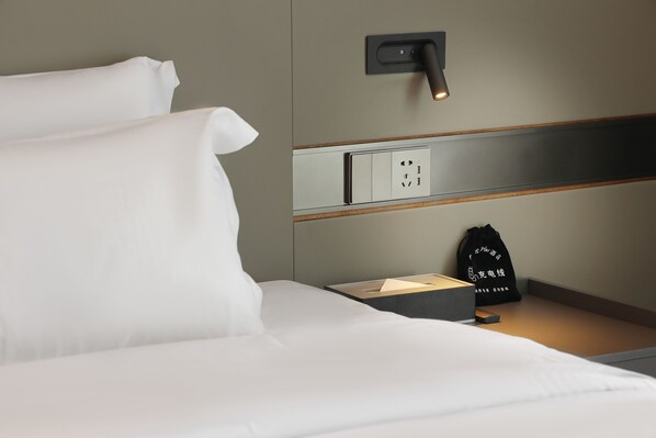 A neatly made bed with white linens, a bedside table with a black bag and a box, and a wall-mounted power outlet. - mantuolunPlusjiudian (Jiangmen)