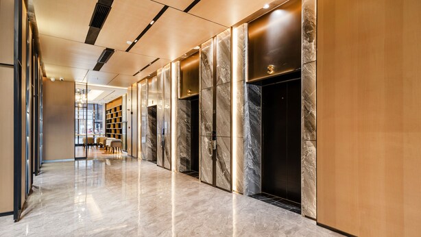 Un pasillo moderno con piso de mármol, un mostrador de recepción de madera y una puerta de cristal que da a un área de descanso.