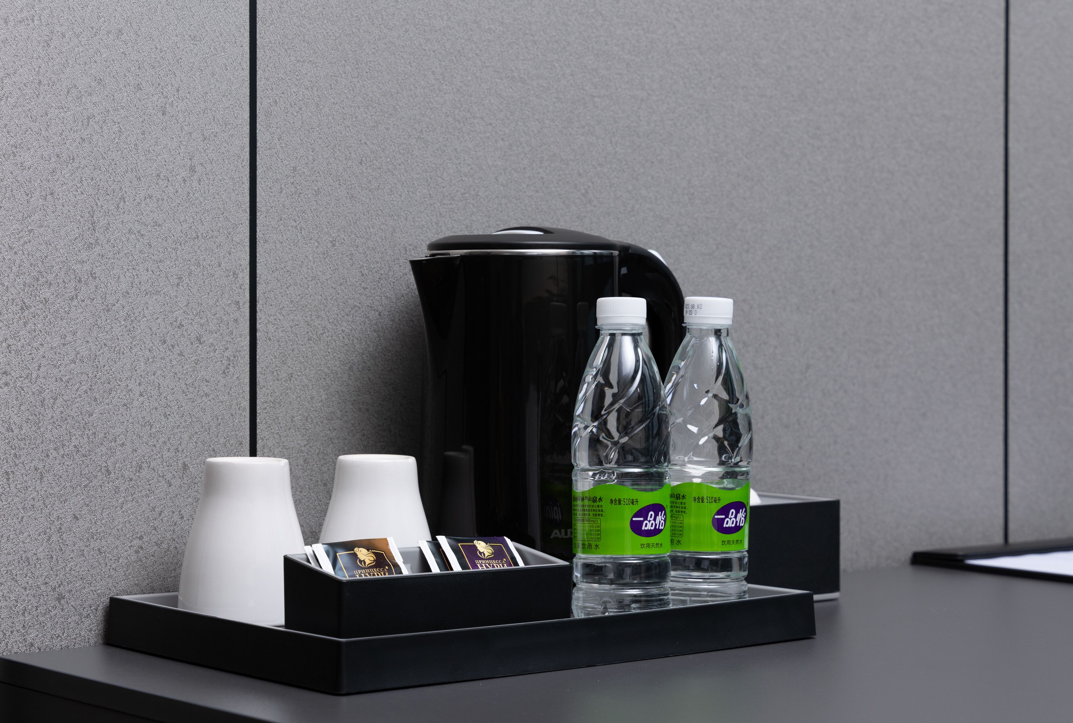 Una cafetera negra, dos lámparas blancas y dos botellas de agua sobre una bandeja negra frente a una pared gris.