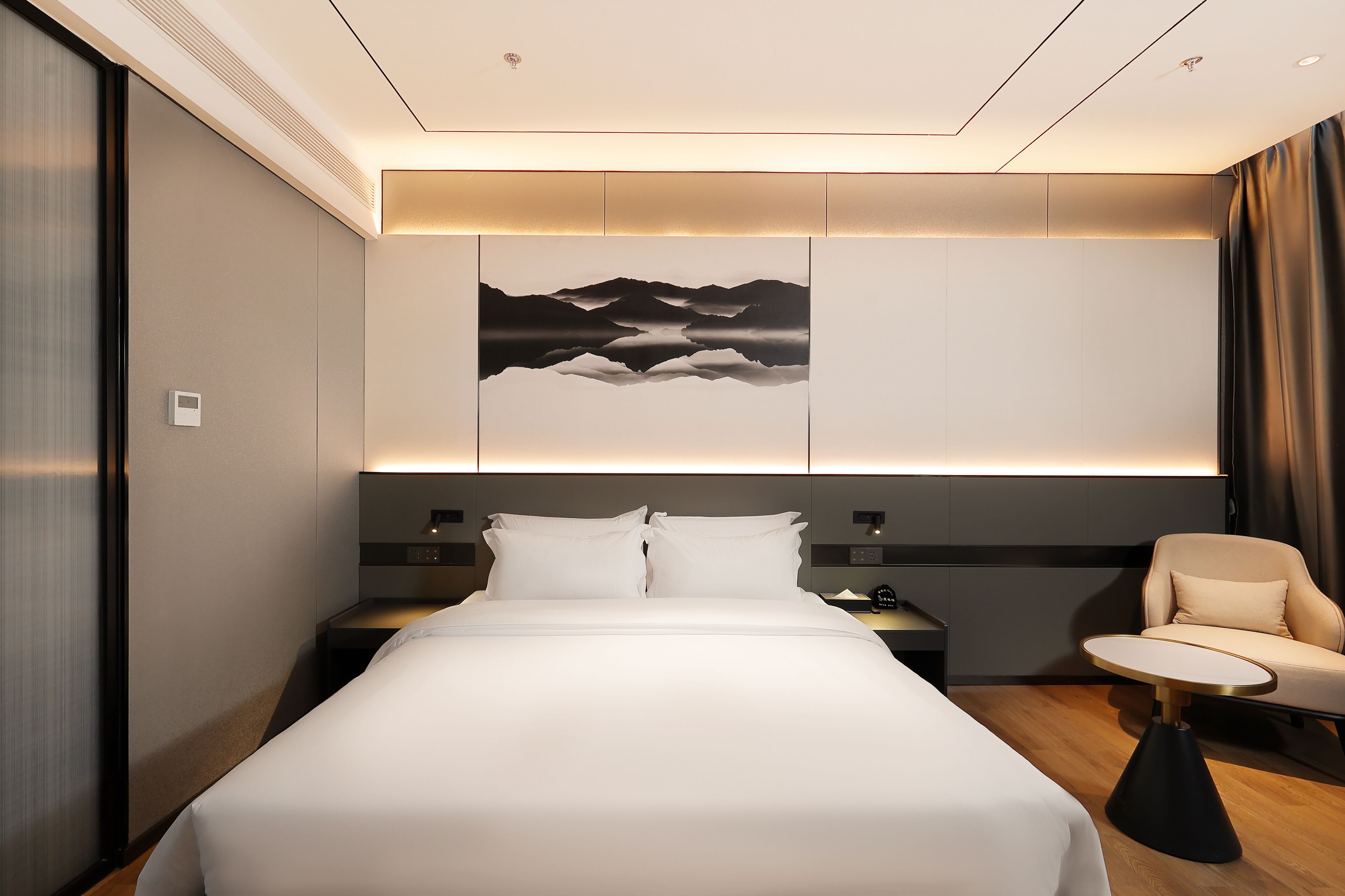 A modern hotel room with a large bed, a bedside table, a chair, and a wall-mounted artwork.