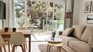 A modern living room with a sofa, a dining table with chairs, a coffee table, and a view of the outdoors through large glass doors. - Sea apartment 1A with pool, 2 min. canyamel beach (Capdepera)