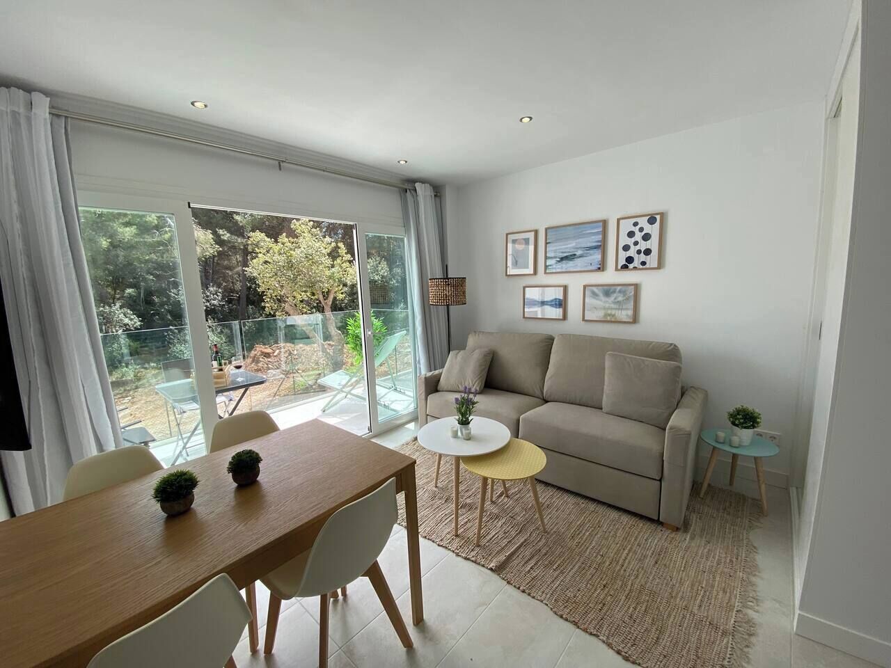 A modern living room with a sofa, a dining table, and chairs. There is a small round table with a plant. The room has large windows and a view of the outdoors.