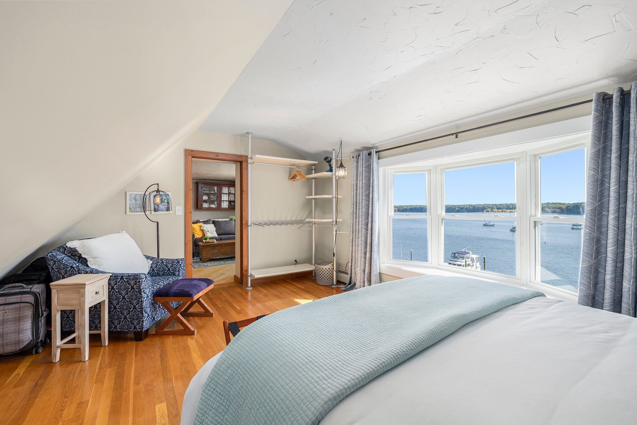 Une chambre avec un grand lit, un bureau, une chaise, une petite table, une étagère et une fenêtre donnant sur la mer.