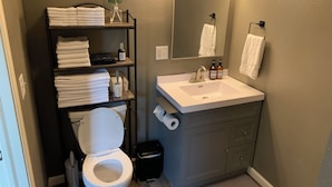 A modern bathroom with a white toilet, a sink with a mirror above it, and a shelf with towels and toiletries.