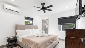 Quarto moderno com cama, mesas de cabeceira, ventilador de teto, ar condicionado, cômoda de madeira e janela com vista para vegetação.