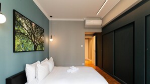 Un lit bien fait, avec du linge de lit blanc et des oreillers, une grande œuvre d’art encadrée représentant un arbre accrochée au mur, et un climatiseur moderne placé au-dessus du lit.