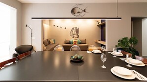 Un espace de repas moderne avec une longue table dressée pour un repas, un élément décoratif mural au-dessus de la table et un coin salon confortable à l’arrière-plan.