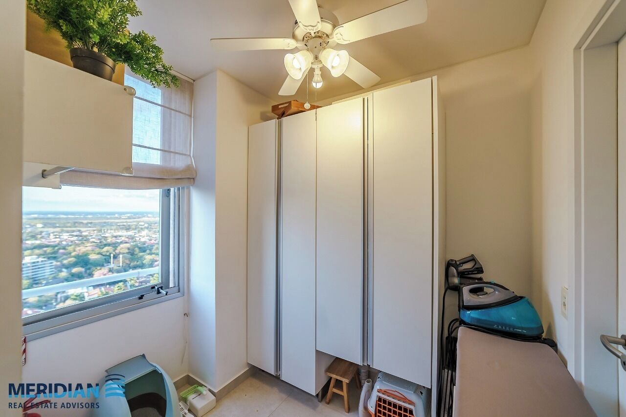 Une chambre dotée d’une grande armoire, d’un ventilateur de plafond, d’une fenêtre donnant sur un paysage urbain et d’un petit tabouret.
