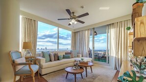 Uma sala de estar moderna com um sofá, duas mesas de centro, uma poltrona e um ventilador de teto. Janelas amplas oferecem vista da cidade.
