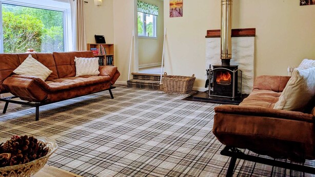 A cozy living room with a brown leather sofa, a plaid rug, a fireplace, and a large window offering a view of greenery. - Holiday cottage tummel valley beautiful views. Own garden dog friendly  (Pershire)